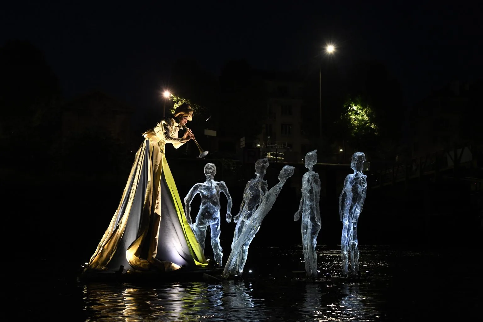 An image of the water theater show Drift by the ilotopie company : The flute player standing on water on a estale foot plays music to transparent and luminous floating human shells.
