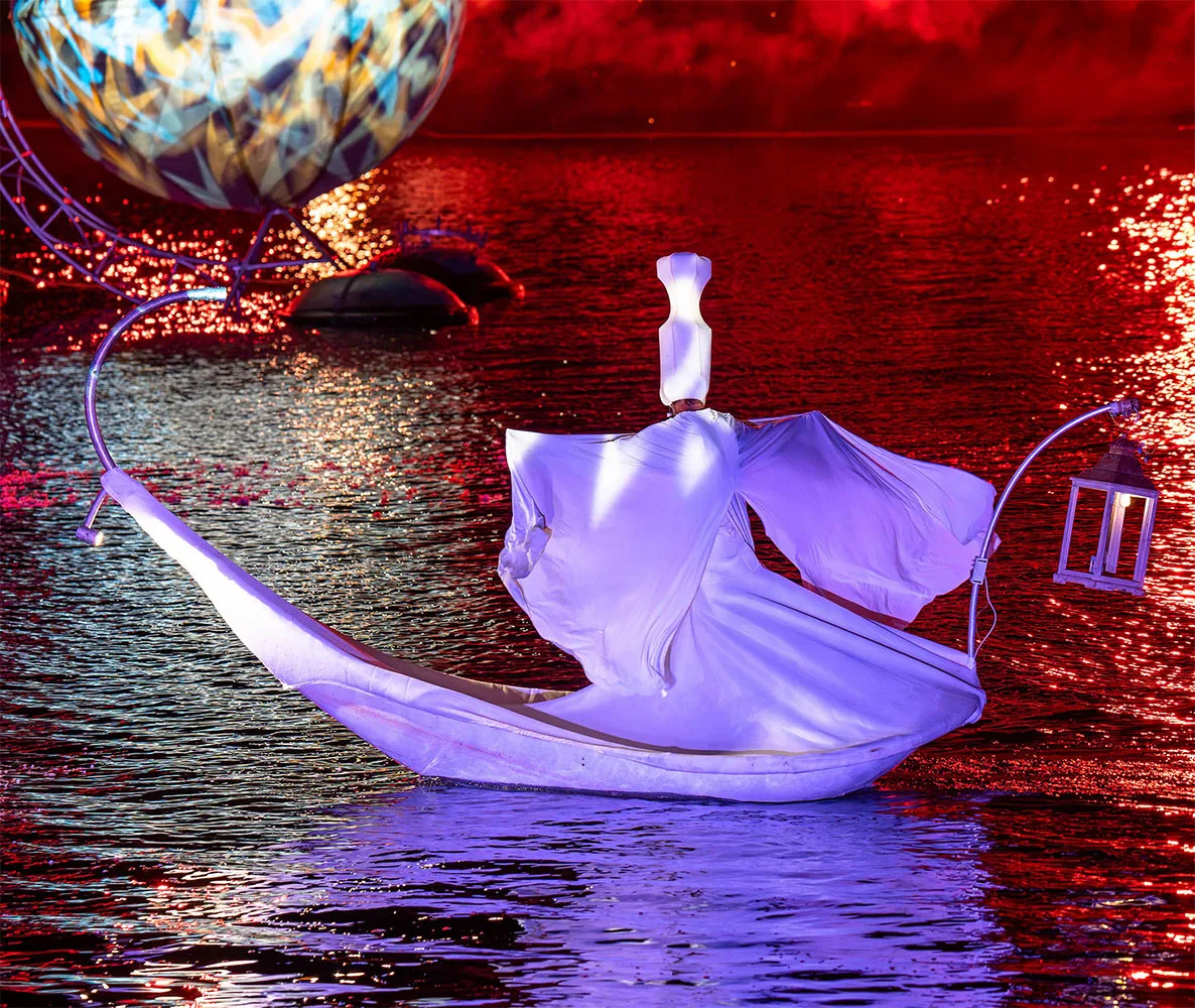 A figure made of white veil in a petal floats in the middle of water which seems to be on fire. in the distance a sphere with kaleidoscope patterns. A moment from the show The Extraordinary Journey of the ilotopie company.