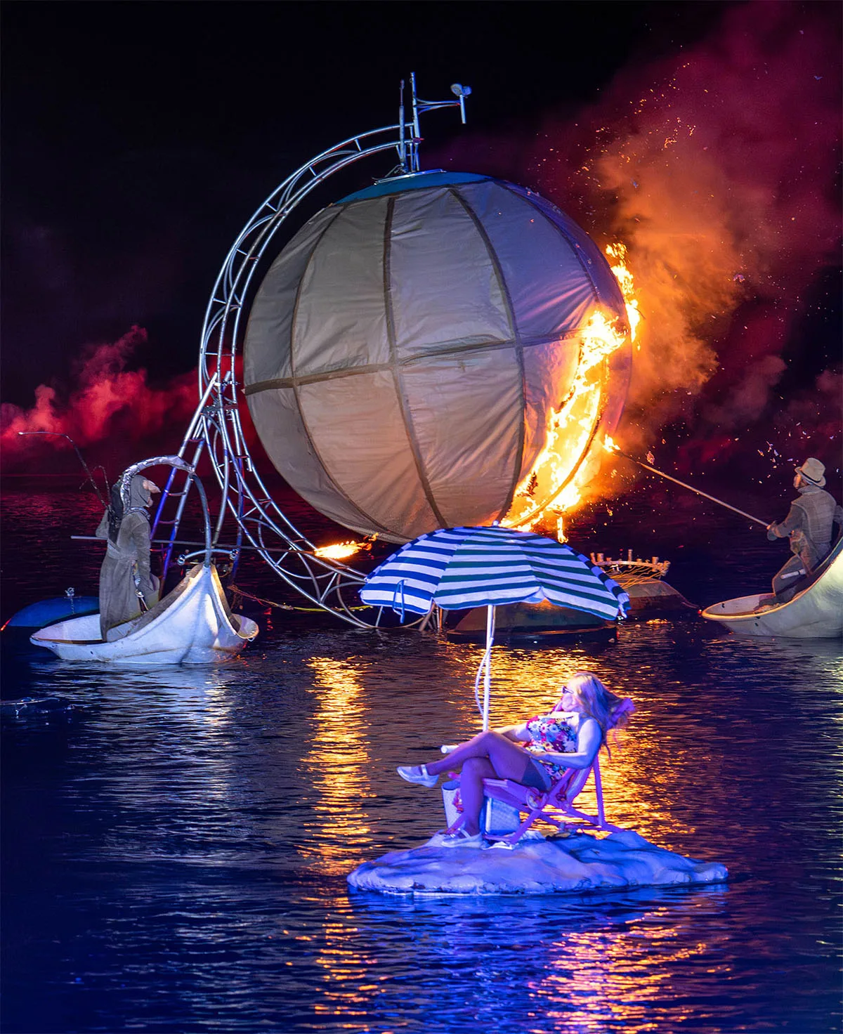 Two devils light a giant sphere on the water, in the foreground a tourist on her personal floating island. One moment from the ilotopie company, the extraodinary journey.