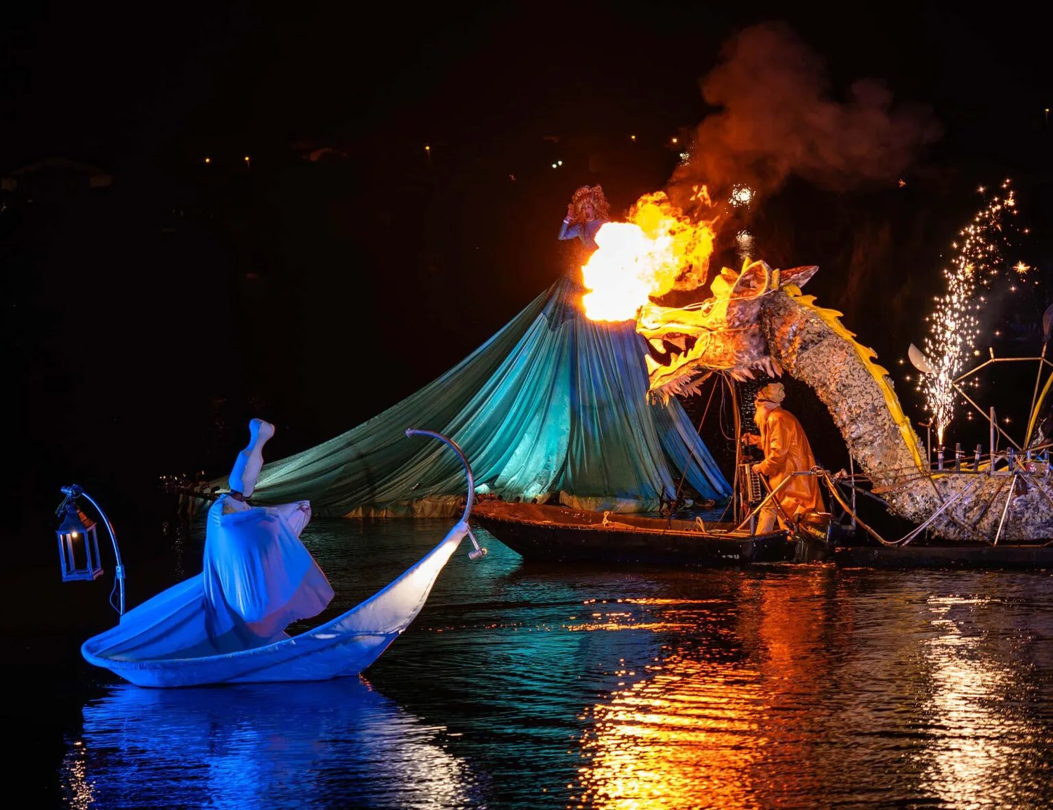 A lady in a big blue dress, a dragon passes in front of her breathing fire. In front of the scene a character all in white in a leaf boat. Everything happens on a body of water which reflects them. It is an event of the show on the water the Extraordinary Journey by ilotopie company.